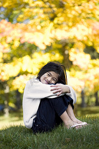 抱膝的小女孩坐在草坪上，背景是明亮的秋叶