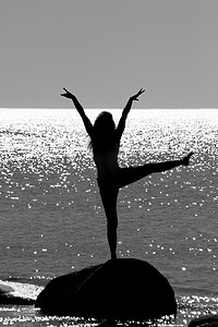 海中石头摄影照片_一个女孩站在海中的一块石头上，双臂和双腿举起的剪影。
