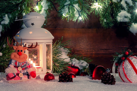 雪地上放着燃烧着蜡烛的白色灯笼，周围环绕着木墙、圣诞树枝和灯的圣诞装饰