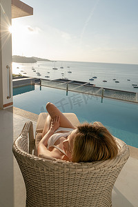 女人躺在阳台上的躺椅上，可欣赏泳池和海边景色。