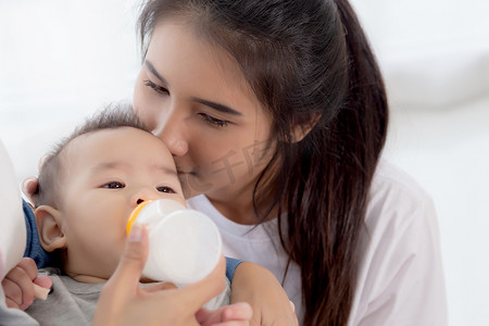 年轻的亚洲母亲在家里用奶瓶拥抱和喂养小女婴，新生儿的纯真与妈妈满意地喝酒，妈妈和孩子的关系和结合，家庭观念。