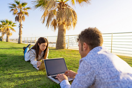 年轻夫妇躺在草地上，在日落或黎明时分，用笔记本电脑面对面地工作。