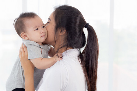 年轻的亚洲母亲在家里的卧室里抱着小女婴，妈妈带着孩子和照顾孩子，女人和孩子无忧无虑，蹒跚学步的孩子和父母，情感和表达，家庭观念。