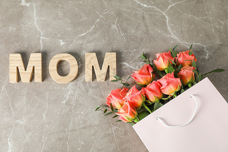 花束妈妈摄影照片_带橙色玫瑰花束的礼品袋和棕色背景上的妈妈铭文，顶视图