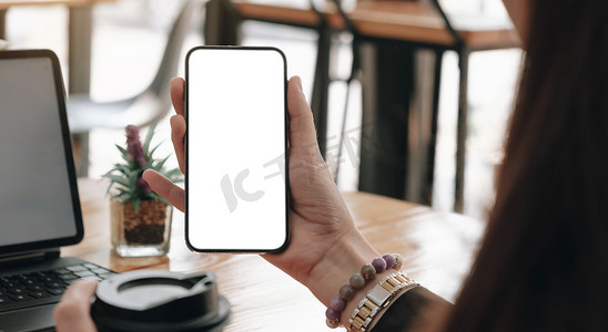 iphonex样机x摄影照片_样机图像，女商人手持空白屏幕移动智能手机，在现代办公室的桌子上使用笔记本电脑和数字平板电脑进行网站或应用程序设计。