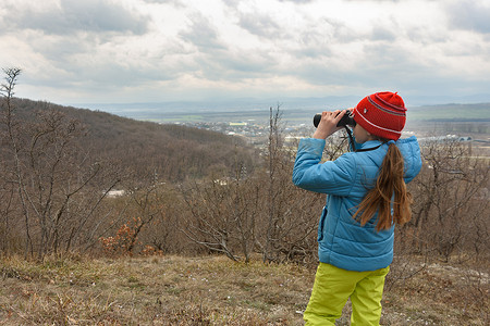 一个女孩通过双筒望远镜检查山景，从后面看