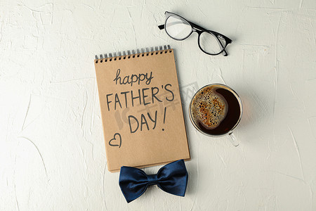 父亲节文字背景摄影照片_笔记本上刻有父亲节快乐、蓝色领结、白色背景的咖啡和眼镜、文字空间和顶视图