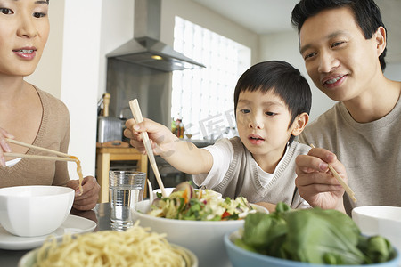 空闲时间摄影照片_年轻的父母看着儿子在餐桌上试图用筷子