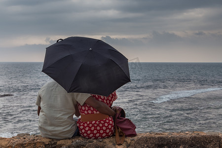 印度夫妇背影坐在海边，撑着伞看着地平线。