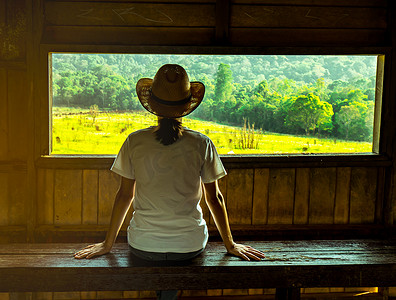 年轻的亚洲女性戴着这顶帽子，坐在木凳上，晚上在野生动物观察塔观看绿草地和森林的美丽景色，阳光温暖。