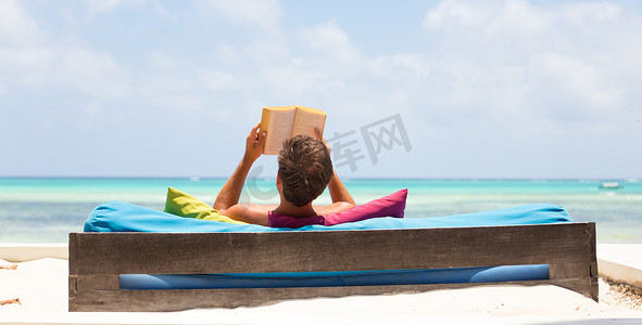 放松的人在豪华躺椅上看书，在美丽的海滩上享受暑假。