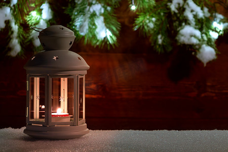 树枝灯笼摄影照片_灯笼在雪地上点燃蜡烛，背景是用圣诞树枝和圣诞灯饰装饰的旧木墙。