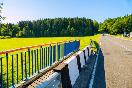 桥的尽头，车子的柏油路在蔚蓝万里的天空下穿过茂密的绿色针叶林，穿过阳光普照的草地。