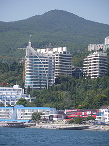 多层现代住宅和酒店在海边的绿树之间，以群山和湛蓝无云的天空为背景。