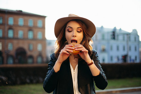戴着帽子的时尚黑发女孩吃美味的汉堡包。