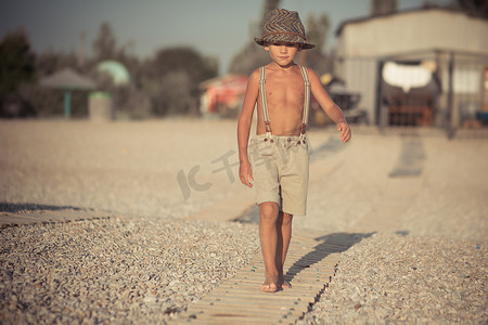 可爱的老式男孩在沙滩上散步，穿着花哨的短裤，戴着绞刑架和绅士帽，独自在令人敬畏的海边享受夏日时光。童年生活的可爱场景