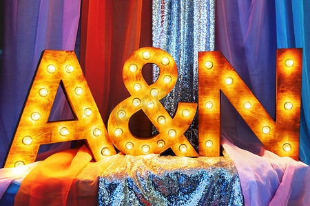 元素趋势摄影照片_婚礼仪式、宴会、派对装饰的时尚设计元素-带有灯泡和闪闪发光的织物的木制字母（新娘和新郎名字中的第一个字母）。