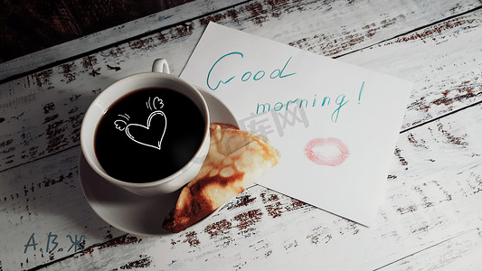 煎饼摄影照片_一杯咖啡，盘子里的煎饼和一张纸上的早安祝福，上面有口红痕迹和滑稽的面孔。