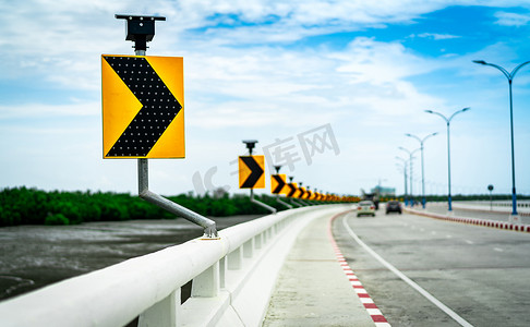 桥上曲线交通标志上的黑色和黄色箭头与太阳能电池板 ob 混凝土道路和汽车的模糊背景靠近泥滩和红树林与美丽的蓝天和云彩。