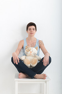 年轻迷人的微笑女性练习瑜伽，坐在半莲花练习 Ardha Padmasana 姿势，穿着深蓝色牛仔裤室内全长，白色毛绒玩具猫在她的腿上锻炼