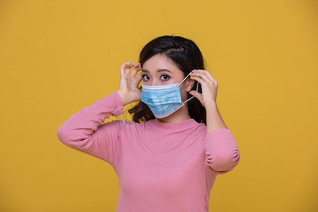 黄色背景下戴着面罩或防护面罩以抵御冠状病毒危机或 COVID-19 爆发的亚洲美丽快乐年轻女性肖像