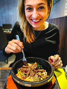韩国石锅拌饭盛在热石锅碗中，被在韩国民族餐厅用餐的白人微笑女性吃掉