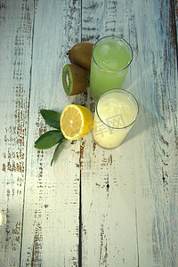 两个玻璃杯，上面放着清爽的果汁和冰块，放在一个纺织品架上，整个和一半切成薄片的橙子，上面有叶子和猕猴桃，放在一张白色的木桌上。