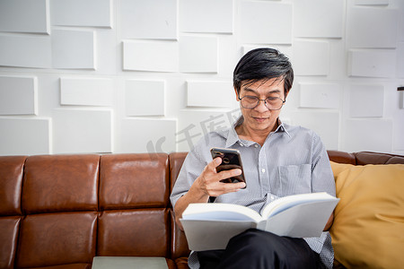 亚洲老人看书，在家里客厅的沙发上使用智能手机亚洲老人肖像是放松和快乐阅读杂志