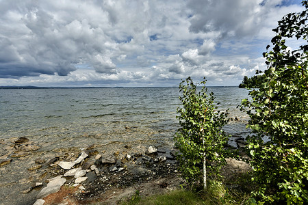 繁茂的树木摄影照片_在湖和多云天空的背景的树木繁茂的岸