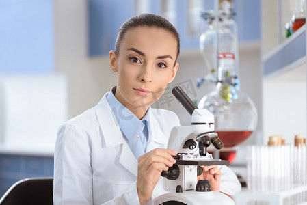 穿着实验室外套的年轻女科学家用显微镜工作，看着相机