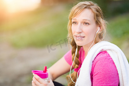 年轻健康的成年女性在户外用毛巾和水瓶穿着健身服，戴着耳机听音乐。