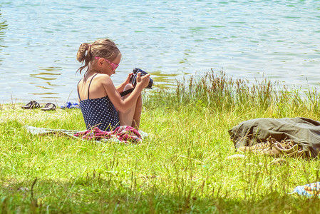 可爱的小女孩在阳光明媚的夏日坐在草地上，用相机拍照。