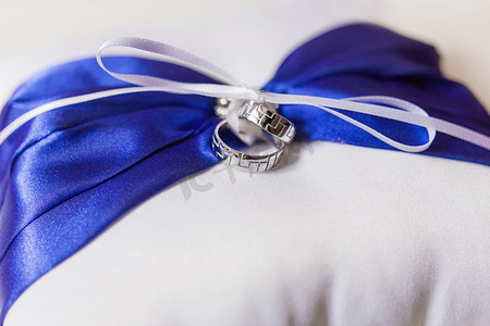一对金色结婚戒指，枕头上有几何装饰，蓝色蝴蝶结和缎带。