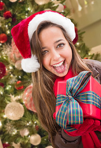 蝴蝶结女孩摄影照片_女孩戴着圣诞圣诞帽，在装饰树前裹着蝴蝶结礼物