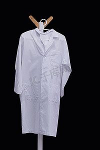白色医生服装摄影照片_白色医用长袍挂在木衣架上，与黑色背景隔离，带有剪裁路径