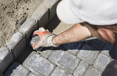 一名建筑工人的手戴着橙色手套，正在铺设新的外部铺路石，小心地将一块铺路石放在水平和倾斜的土壤基础上。