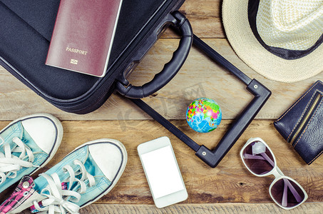着装旅行者的护照、钱包、眼镜、手表、智能手机设备、帽子、鞋子，放在木地板上准备旅行的行李上。
