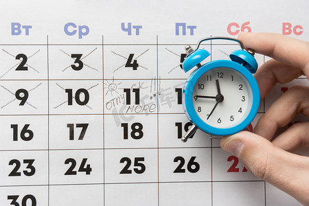手帐背景摄影照片_一只手拿着一个小闹钟，背景是一个日历，上面标有日期和假期