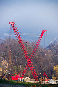 带有红色绞架的建筑起重机的施工过程看起来像字母 X。秋天的雾和河桥与模糊背景下的大型浮动酒店。