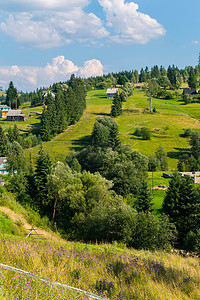绿色山坡上的村屋，在蓝色多云的天空下有夏季田野和高大的树木。