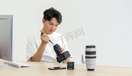 吹风机创意摄影照片_一位亚洲摄影师正在用吹风机清洁相机和镜头，以便在他的办公桌上进行清洁。