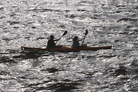 两个人沿着海湾划着皮划艇，在水面上反射出耀眼的阳光。