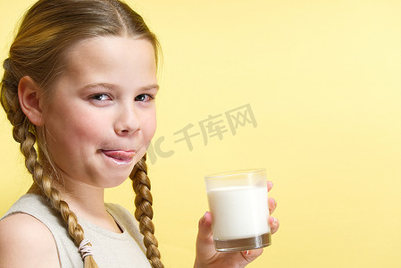 牛奶胡子摄影照片_留着辫子和牛奶胡子的女孩在黄色背景下喝牛奶。