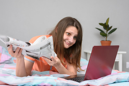一个手里拿着报纸的女孩躺在床上，看着笔记本电脑屏幕寻找工作