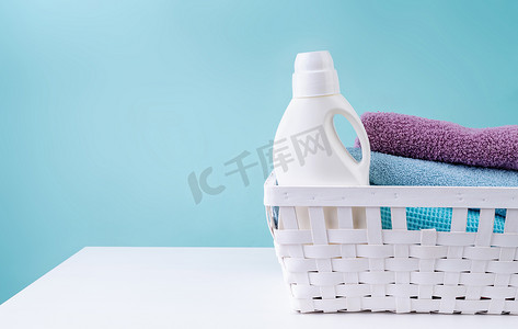 蓝色背景中突显的白桌上带洗涤剂瓶和一堆干净毛巾的洗衣篮
