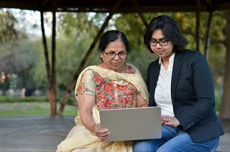 年轻的印度妇女坐在印度新德里的一个公园里，用笔记本电脑帮助她退休的老母亲。