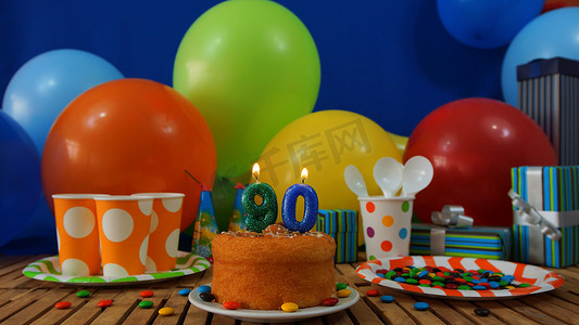 青色摄影照片_在土气木桌上的生日蛋糕有五颜六色的气球、礼物、塑料杯子和塑料盘子的背景有糖果和蓝色墙壁的在背景中