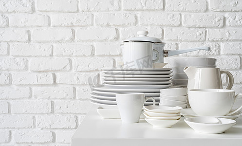 陶瓷器皿摄影照片_白色砖墙背景的桌子上堆放着白色陶瓷盘子和餐具