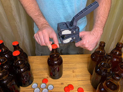 在家里酿造精酿啤酒，男人在木桌上用塑料封盖器关闭棕色玻璃啤酒瓶，上面有红色皇冠盖。