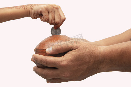 手拿着用泥土制成的印度便士银行，儿童手在孤立的背景下将硬币放入陶罐或陶土存钱罐中 — 储蓄、投资或捐赠的概念展示。